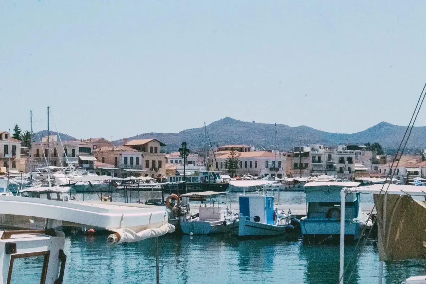 Tranquil Mediterranean harbor scene with boats, mountains, and traditional architecture under clear skies.
