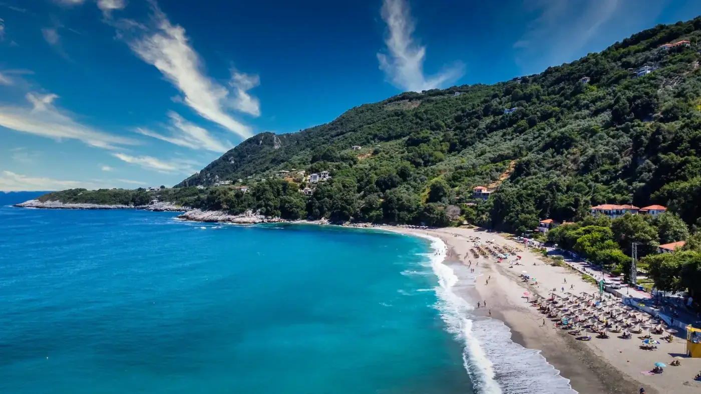 Stunning aerial view of Agios Ioannis beach in Greece, showcasing clear turquoise waters, lush greenery, and mountain backdrop.