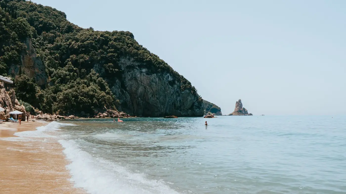 Peaceful beach scene at Agios Gordios with clear blue waters and lush green cliffs in Greece.