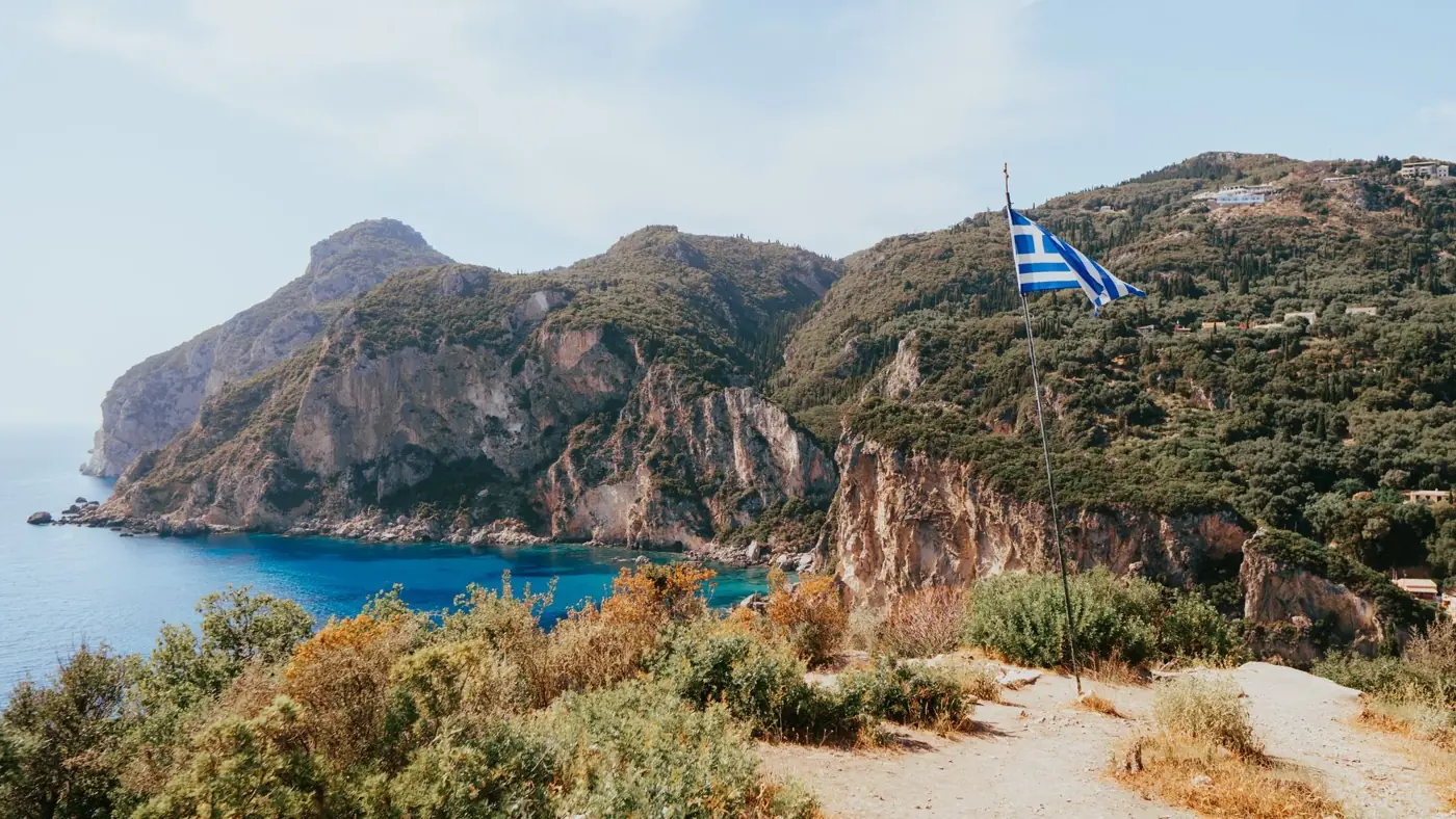 Beautiful view of the Greek flag waving over the cliffs and sea in Corfu, Greece.