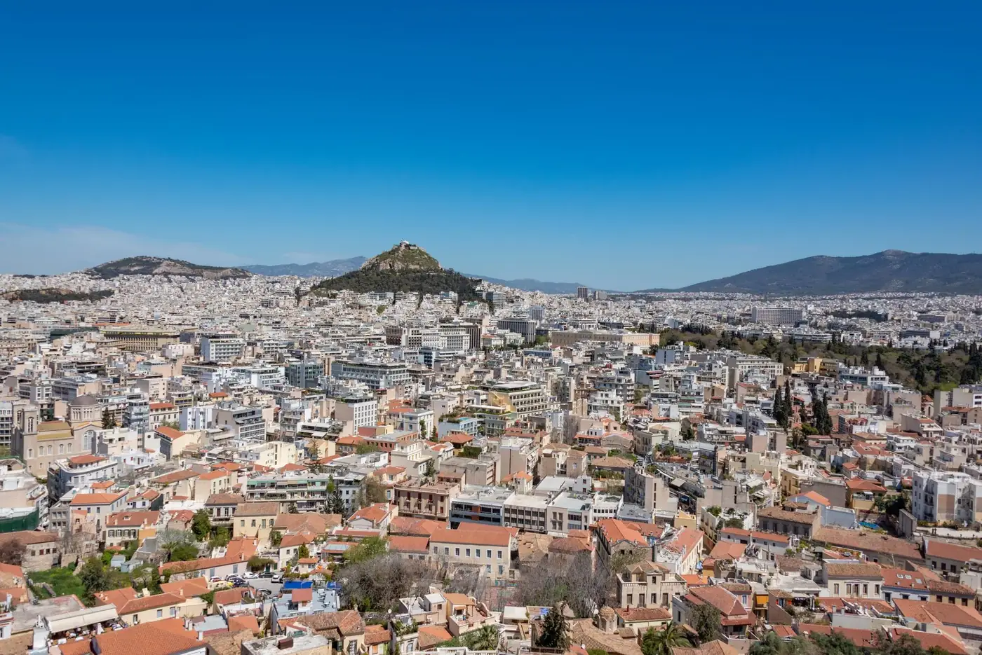 Expansive aerial view of Athens, showcasing the vibrant cityscape and iconic Lycabettus Hill under clear blue skies.