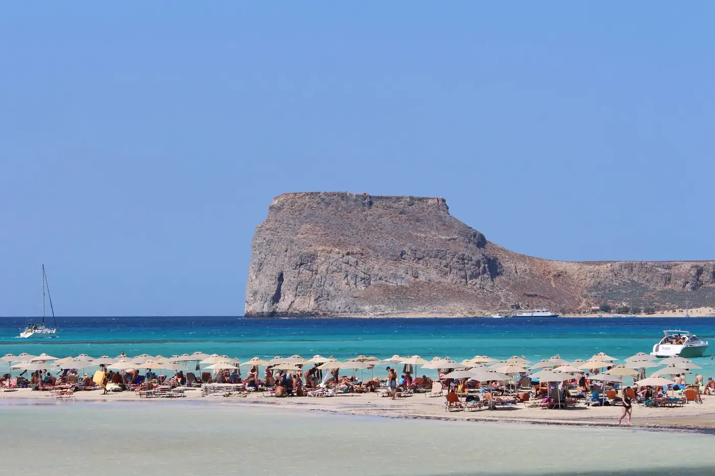Crowd of travelers on sandy shore under umbrellas near azure ocean with rocky mountain in sunny day in tropical lagoon