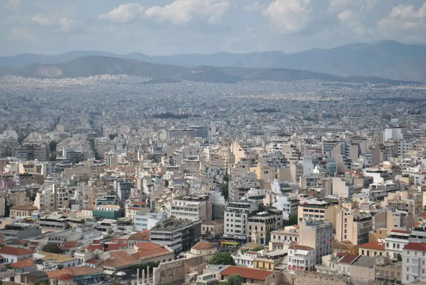 Aerial photo capturing Athens, Greece's sprawling cityscape, with mountains and clear skies in the background.