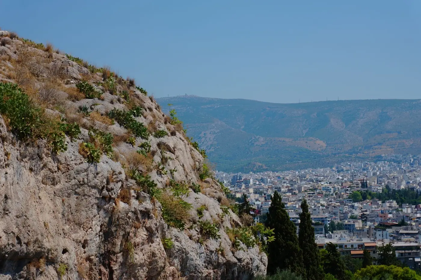 View of Athens cityscape with rocky outcrop and greenery under a bright summer sky.