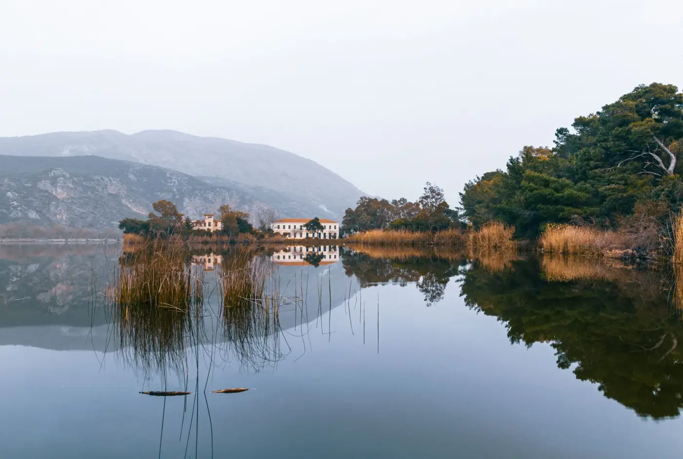 Tranquil autumn scene at Kaiafas Lake with foggy reflections, trees, and mountains.