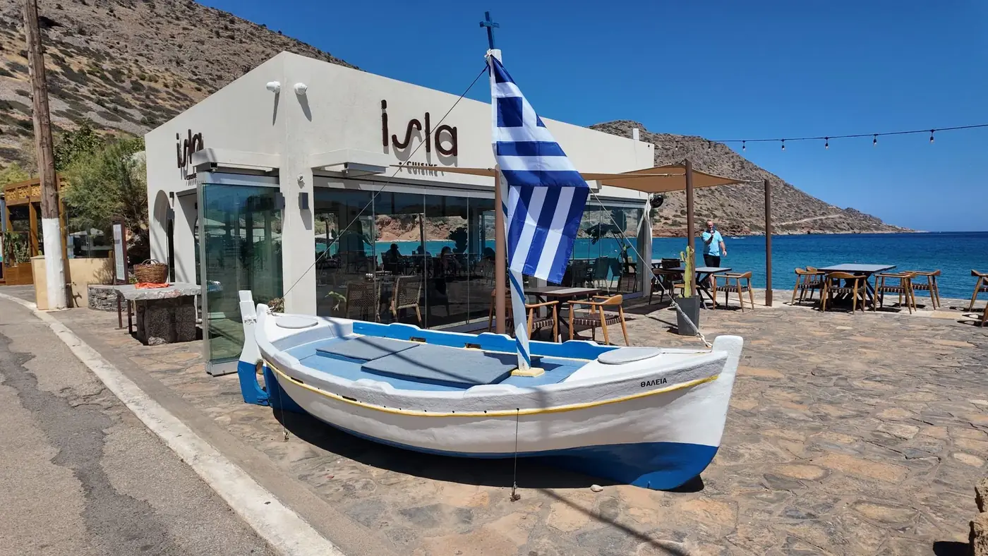 A scenic view of a Greek restaurant by the sea in Plaka with a traditional boat and Greek flag.