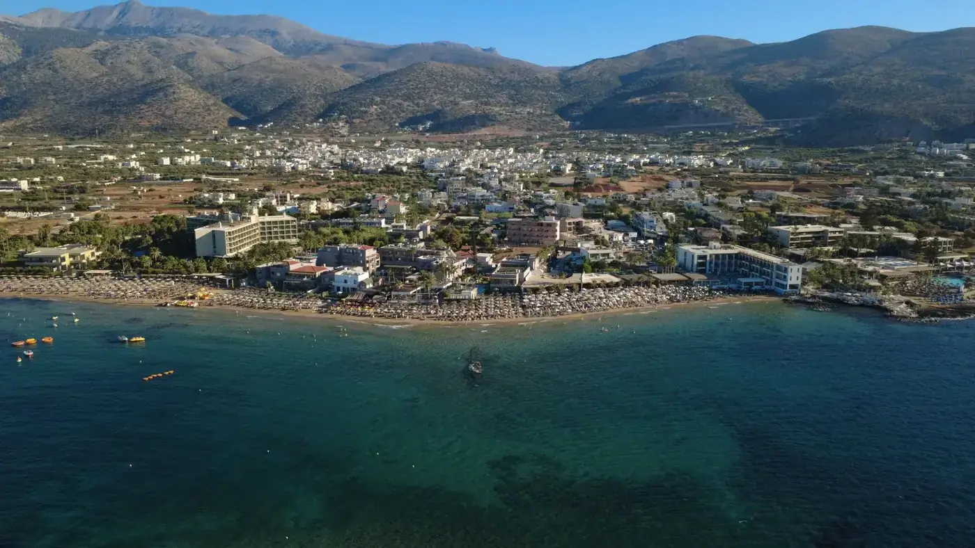 Stunning aerial photo showcasing the beach and town of Malia, Crete, Greece with mountainous backdrop.