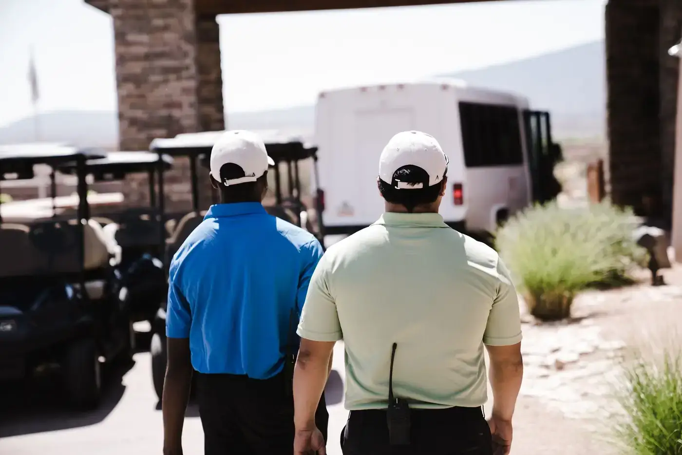 Two men wearing uniforms and caps walking outdoors near golf carts and a van.