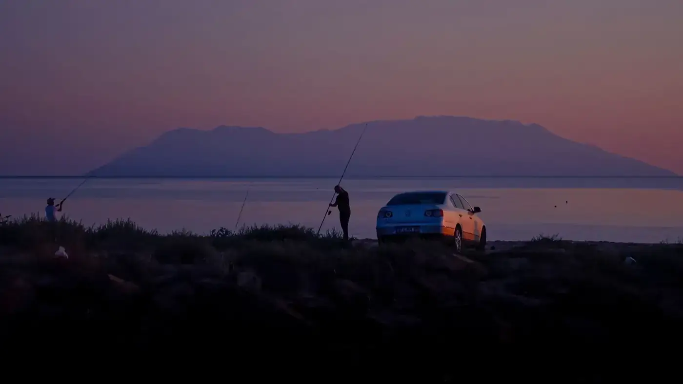 Fishermen casting lines near a car at dusk with a scenic mountain view.