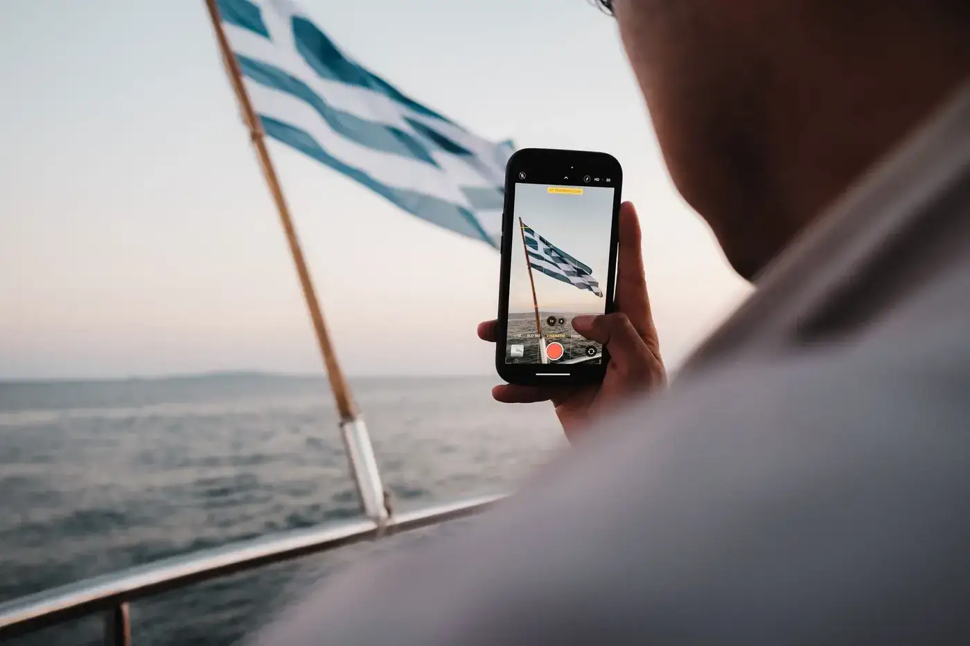 A person captures the Greek flag on a smartphone while sailing on a calm sea during the day.