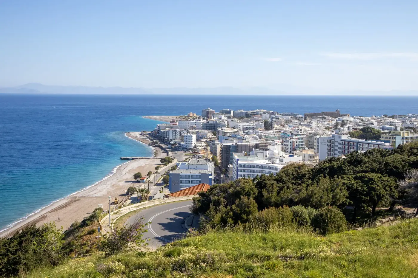 Aerial view of a coastal city with beaches, green hills, and a vast blue sea under a clear sky.