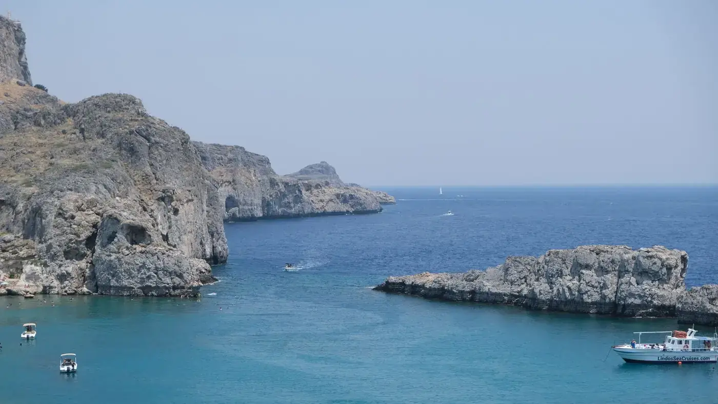Serene view of Lindos coast with clear blue water, rocky cliffs, and boats floating under a clear sky.