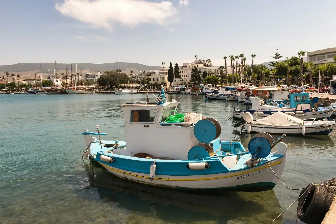 A serene view of fishing boats moored in the picturesque harbor of Kos, Greece, under a bright summer sky.