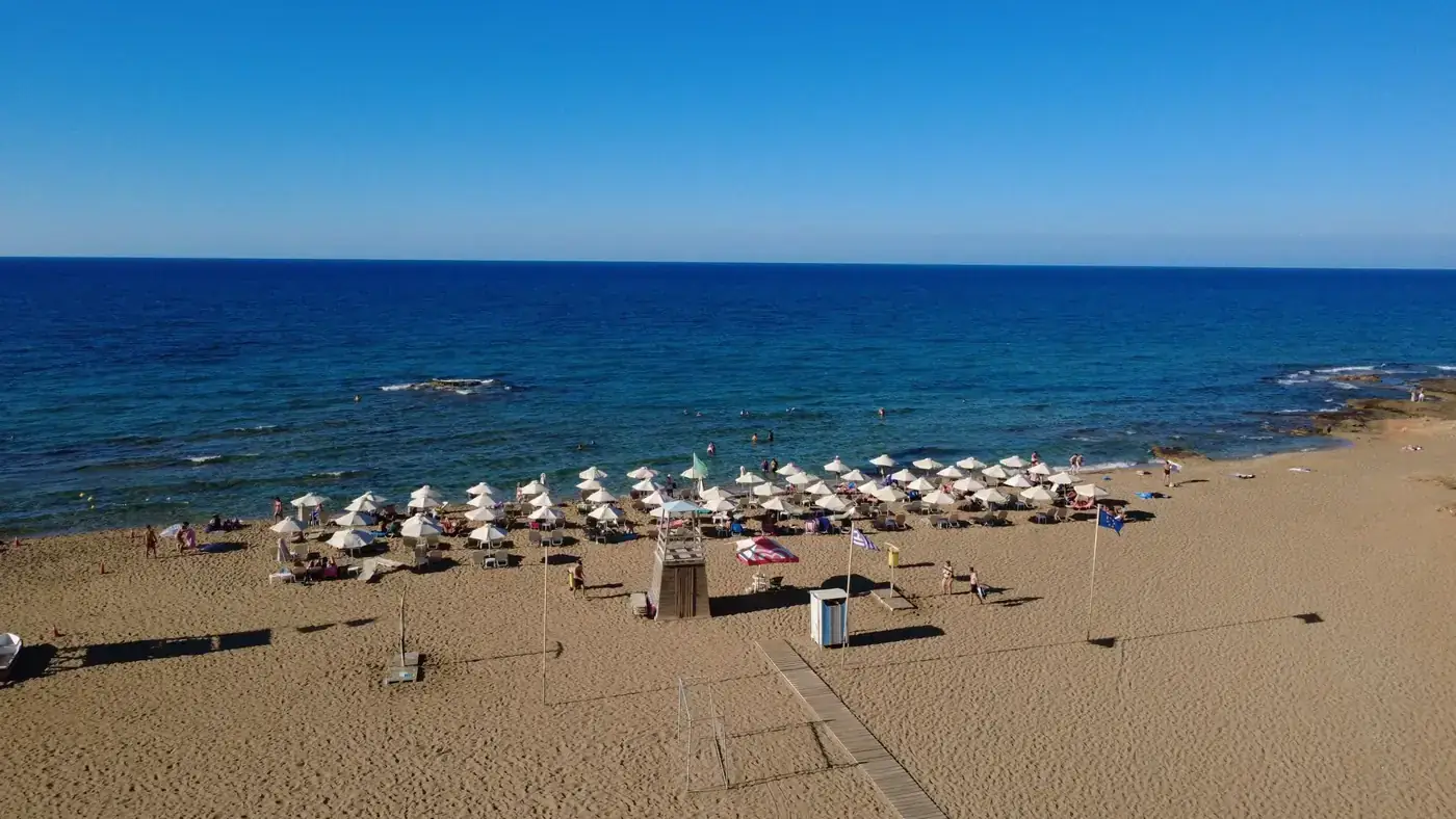 Aerial view of a sunny beach with umbrellas and clear blue sea in Malia, Greece.