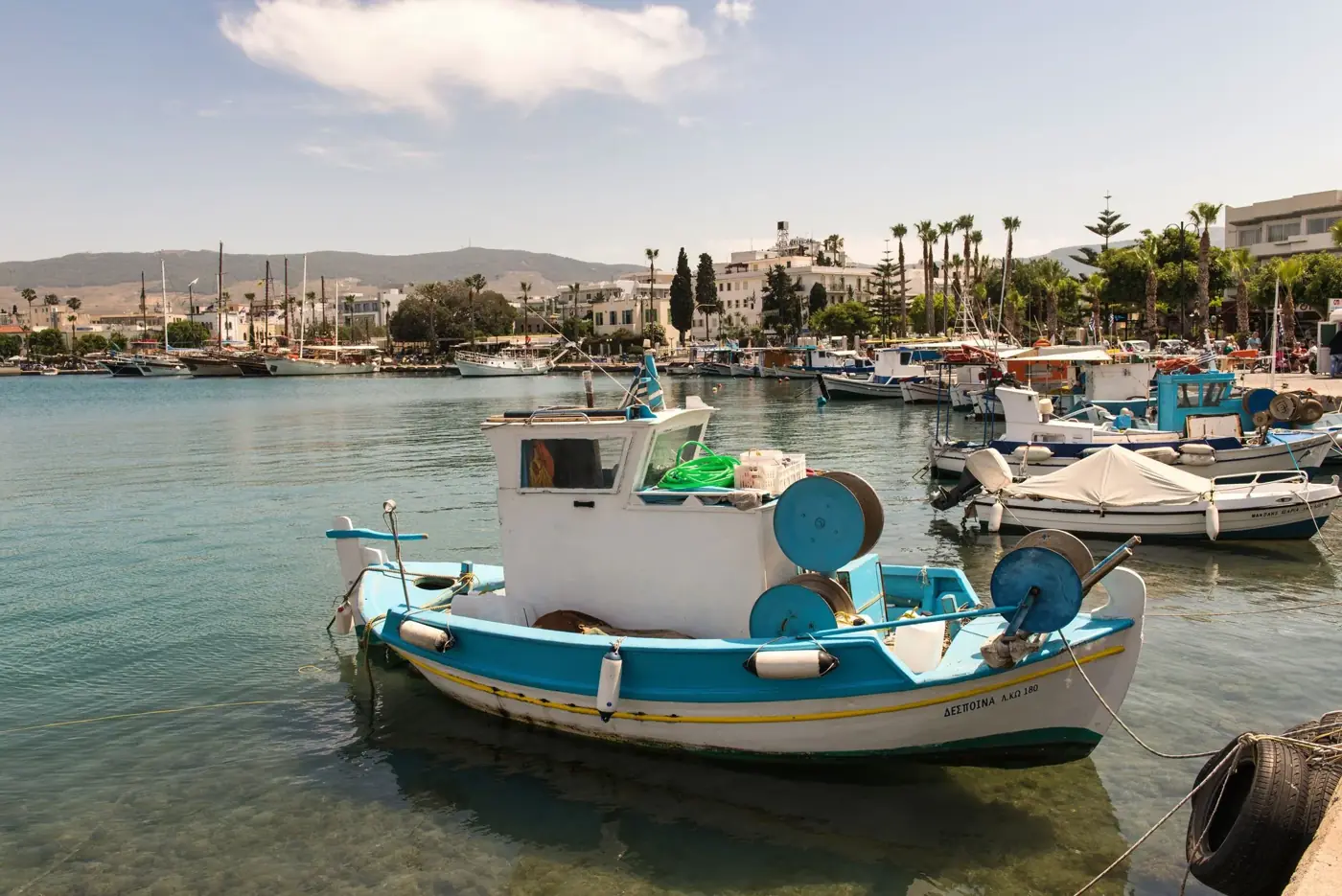 A serene view of fishing boats moored in the picturesque harbor of Kos, Greece, under a bright summer sky.
