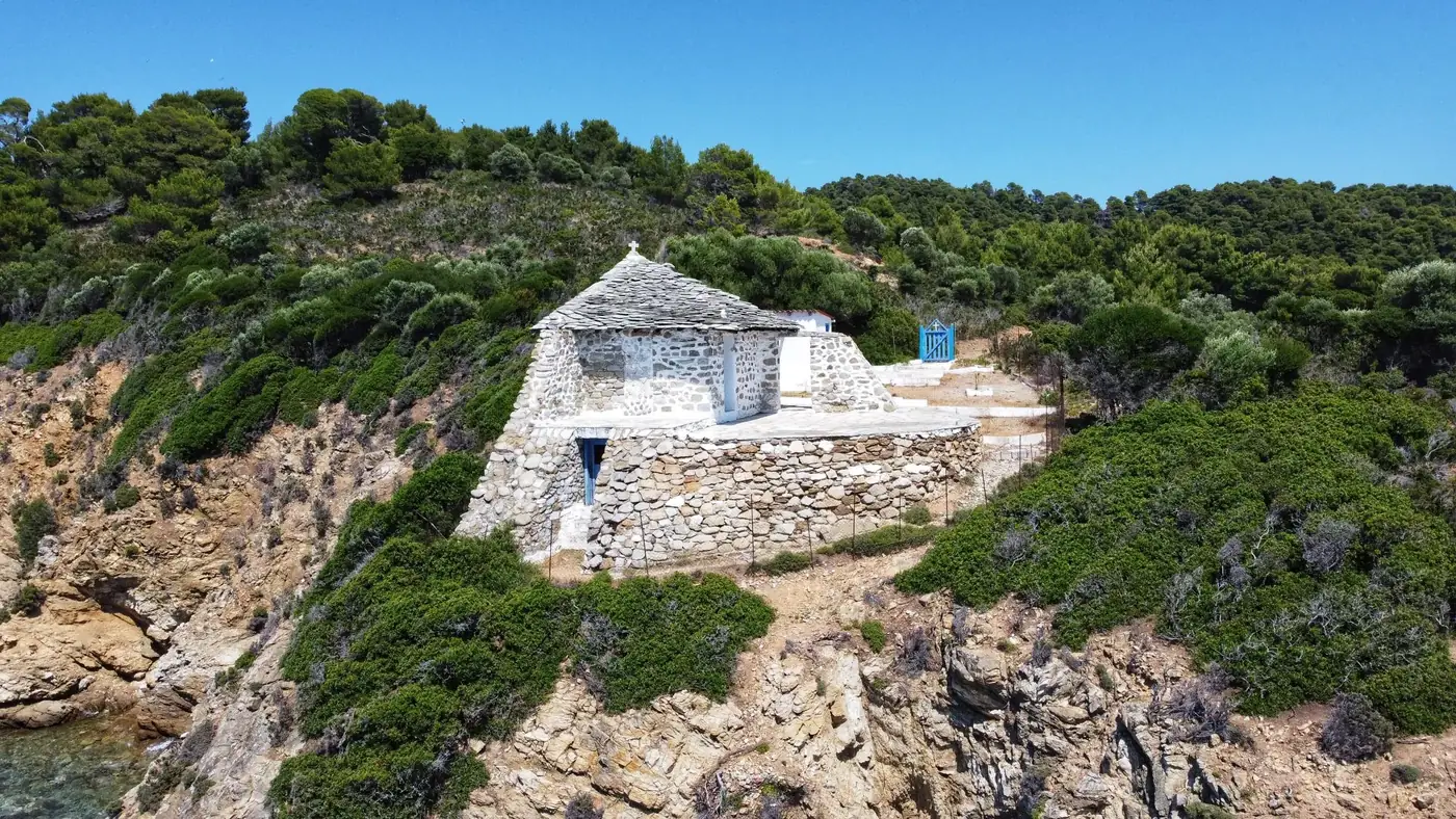 Aerial view of a rustic stone house on a Greek coastline surrounded by lush greenery and clear blue skies.