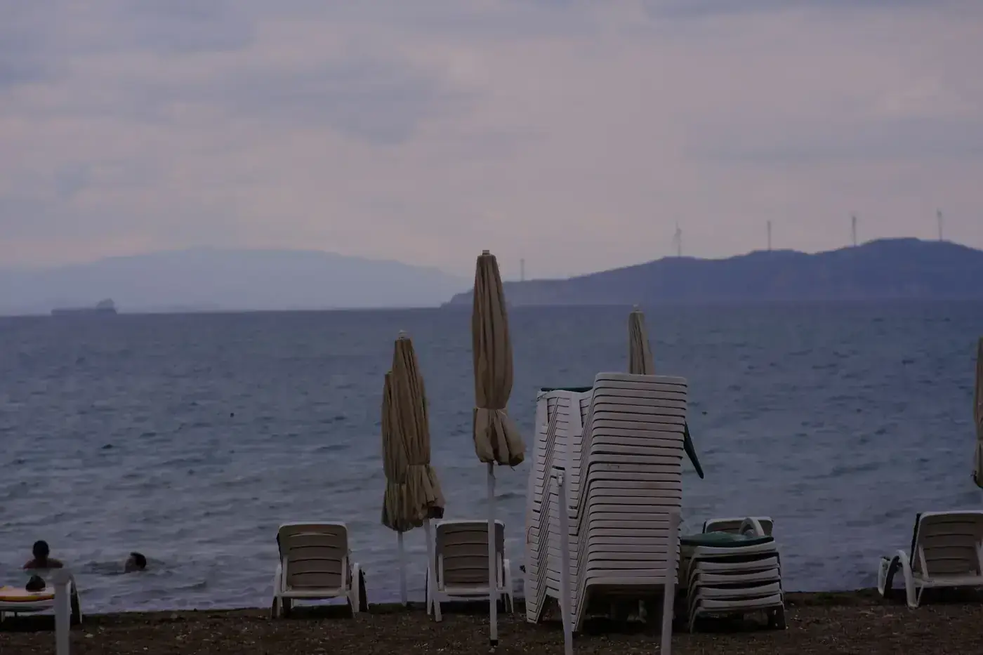 Foça beach scene with empty sunbeds and umbrellas against a cloudy backdrop.