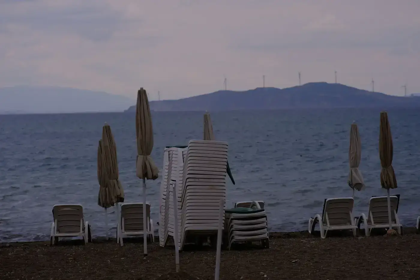 A serene beach in Foça, İzmir with empty loungers framed by distant wind turbines.