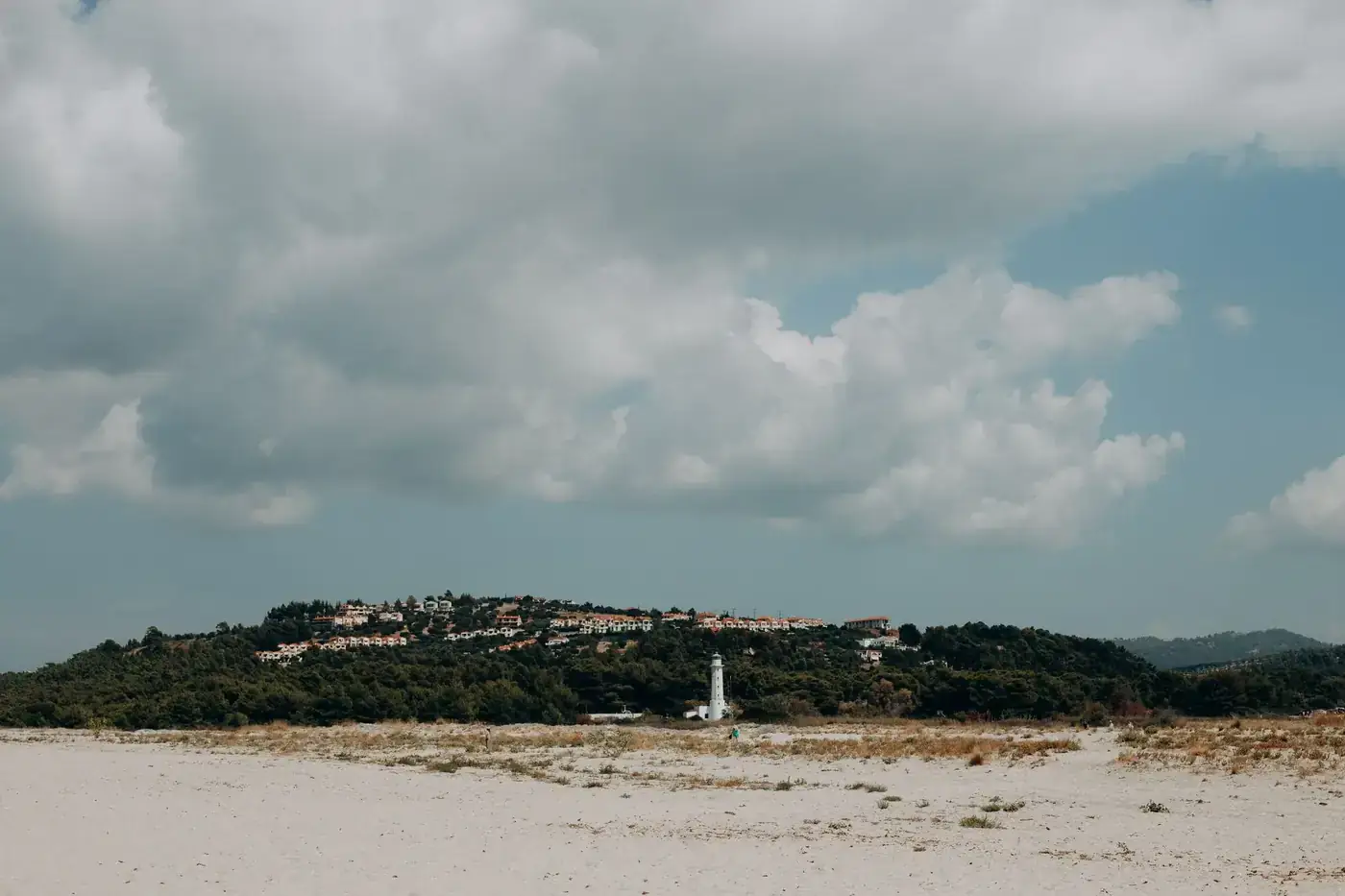 Scenic Greek beach landscape with lighthouse and hillside village under fluffy clouds.