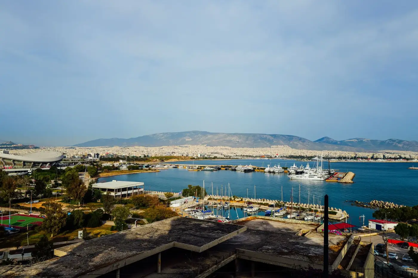 A picturesque marina view with yachts, cityscape, and distant mountains under a clear sky.