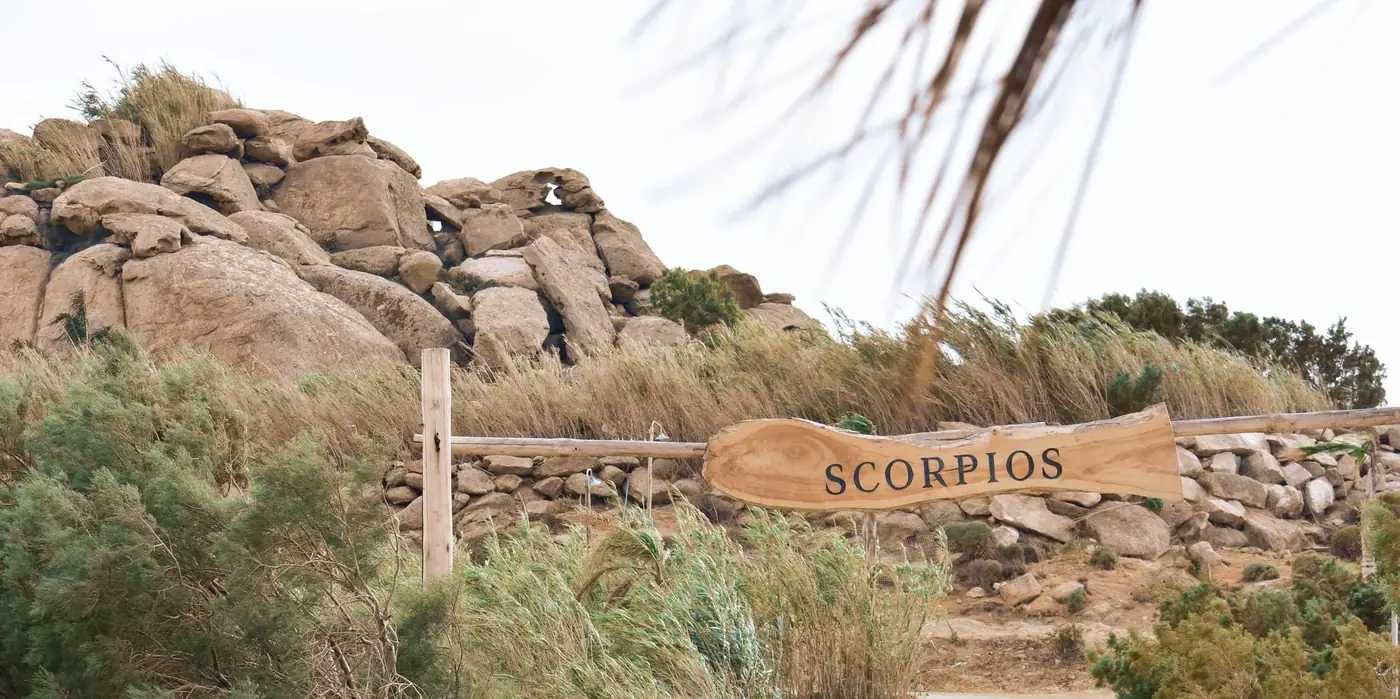 Wooden sign for Scorpios amidst rocky terrain and lush greenery in Mykonos, Greece.