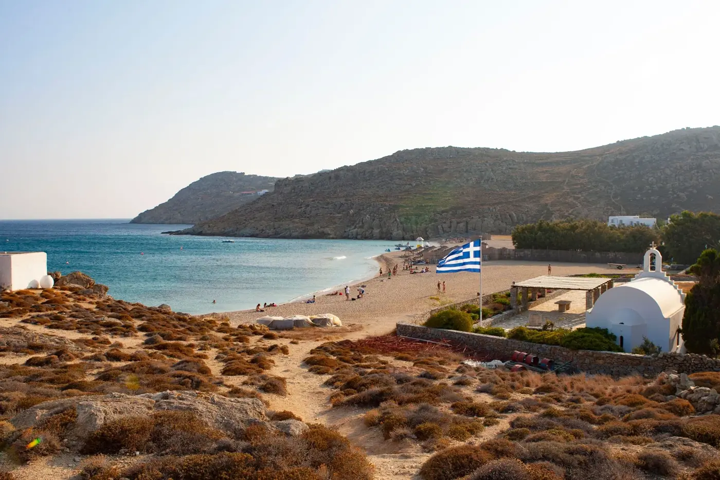 Scenic view of a beach in Mykonos with a Greek flag and traditional church.