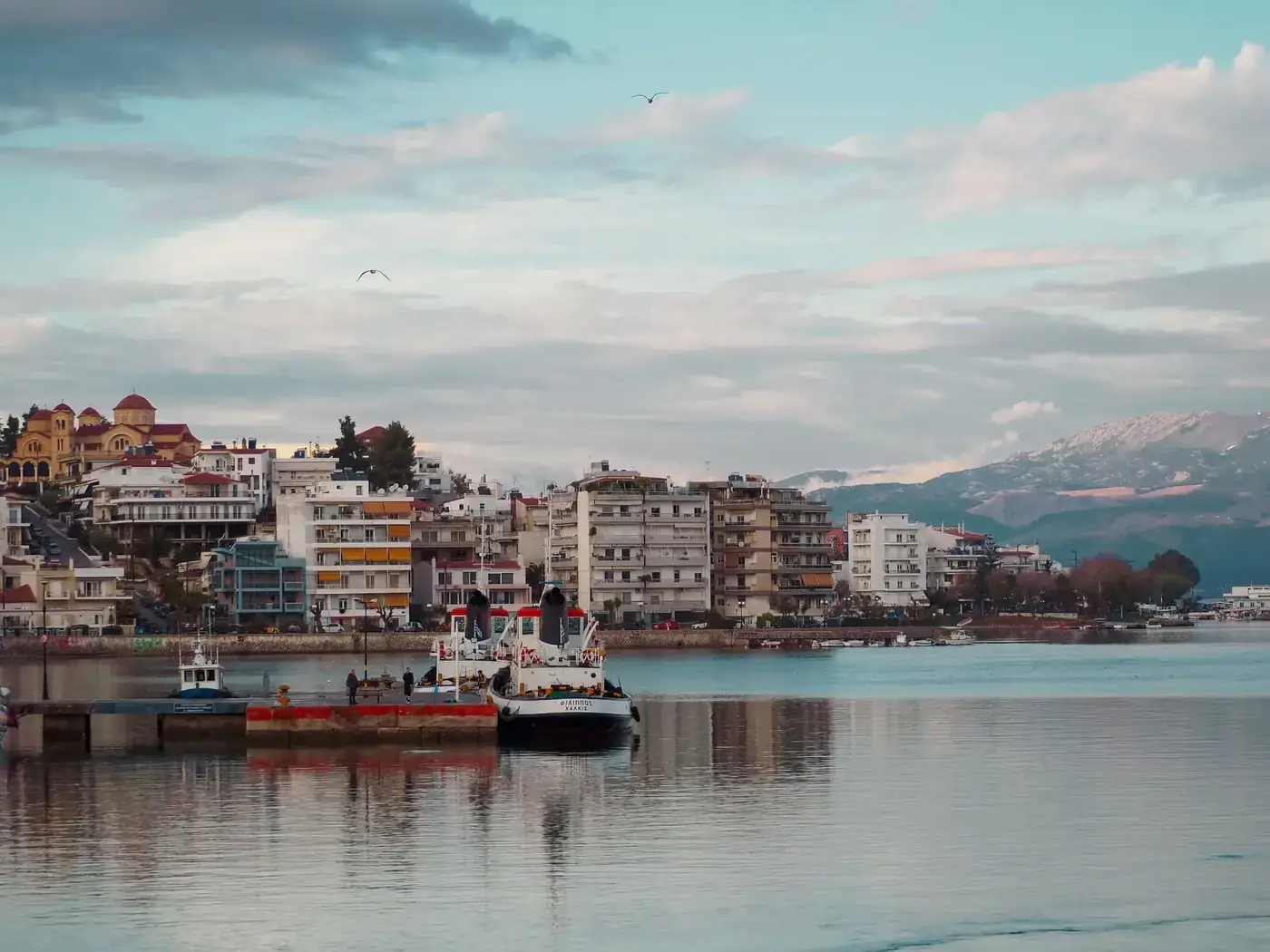 A picturesque waterfront cityscape featuring boats, buildings, and mountains under a scenic sky.