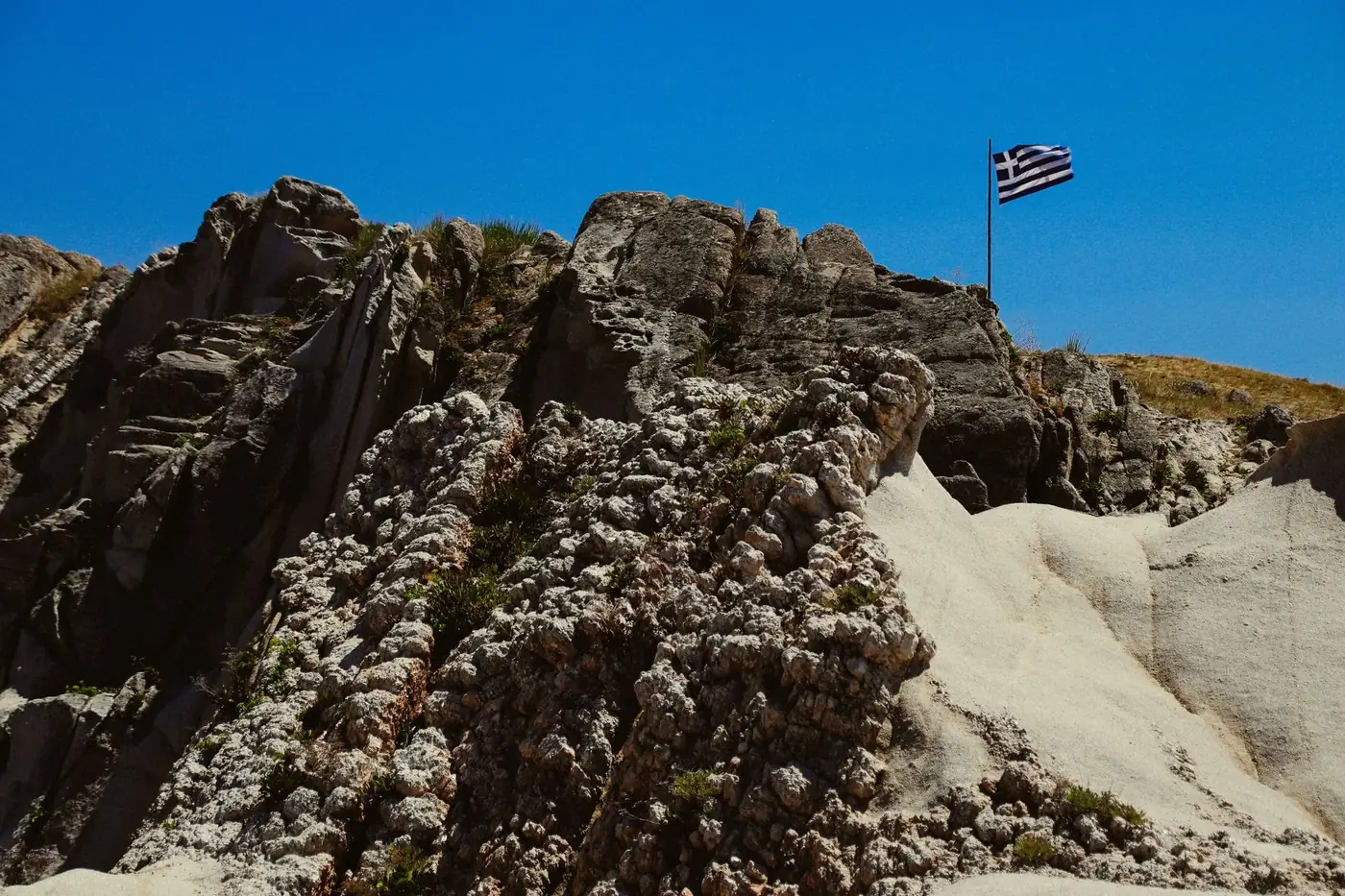 Eroded rocky landscape with Greek flag on a hilltop under bright blue sky in Toroni, Greece.