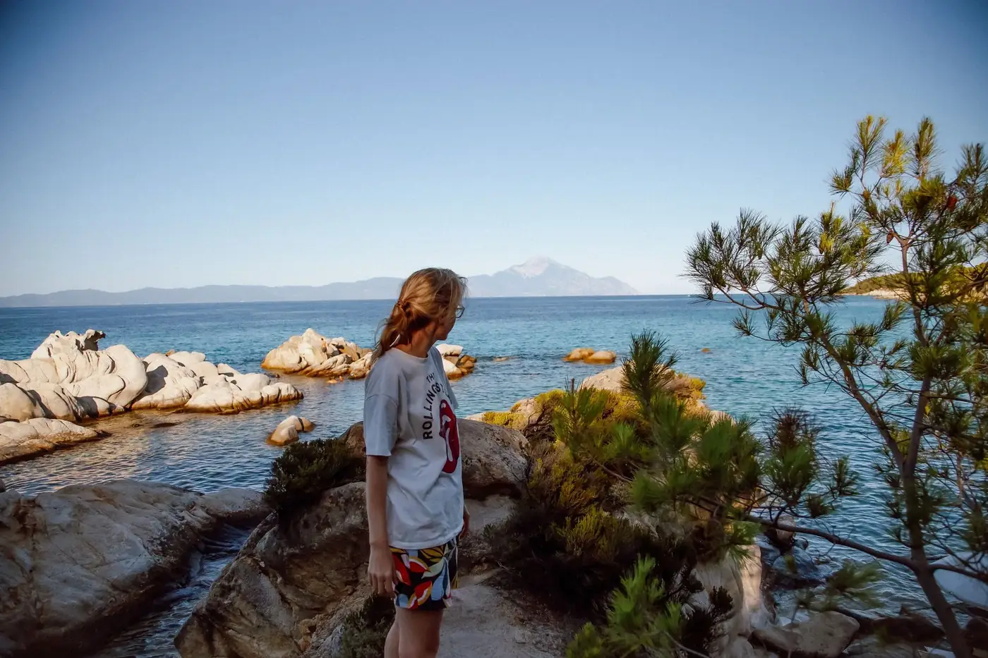 A young woman stands by the rocky Greek coastline, enjoying the summer view.