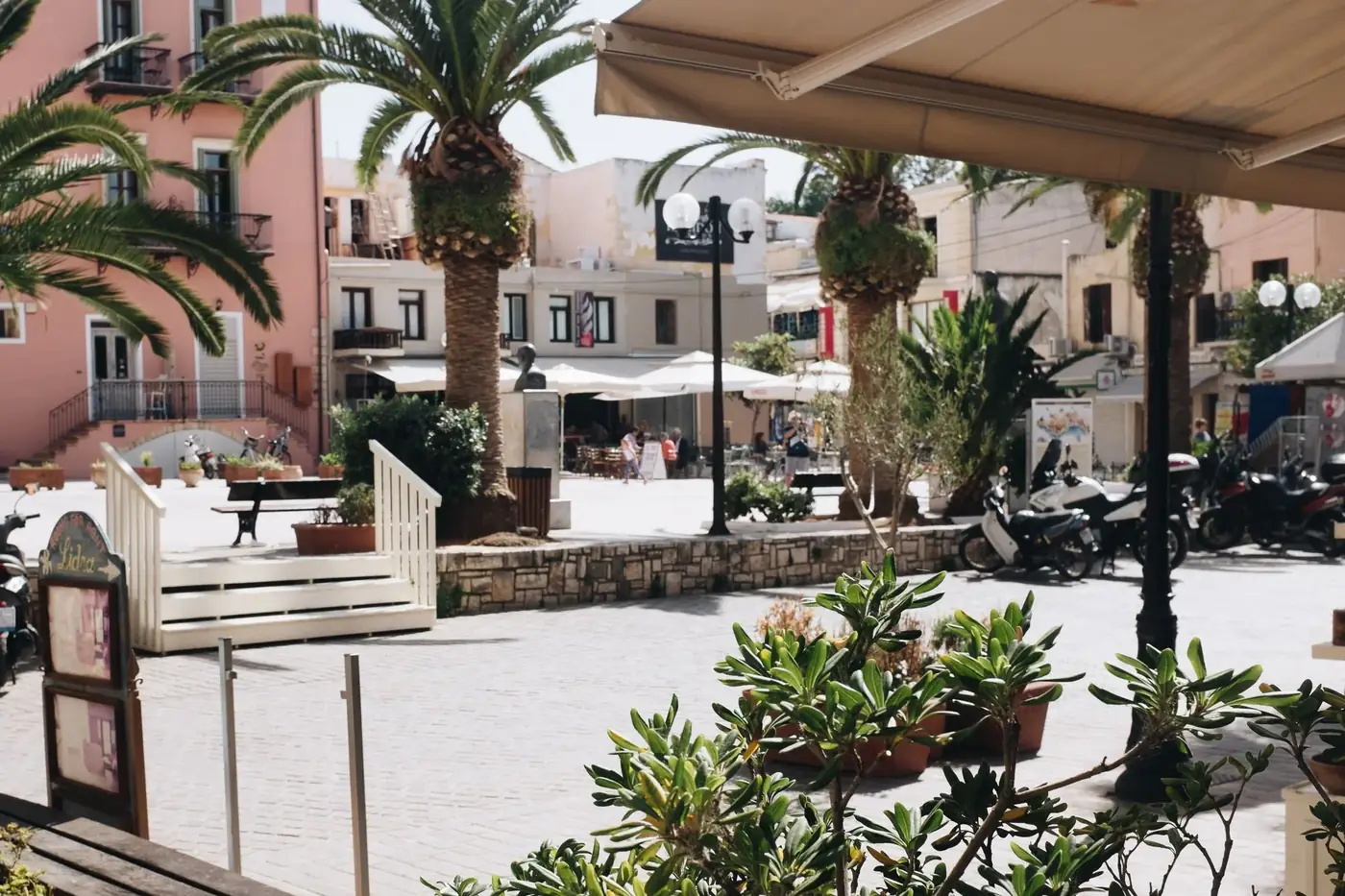 Vibrant square in Chania, Greece, showcasing palm trees, cafes, and charming architecture.