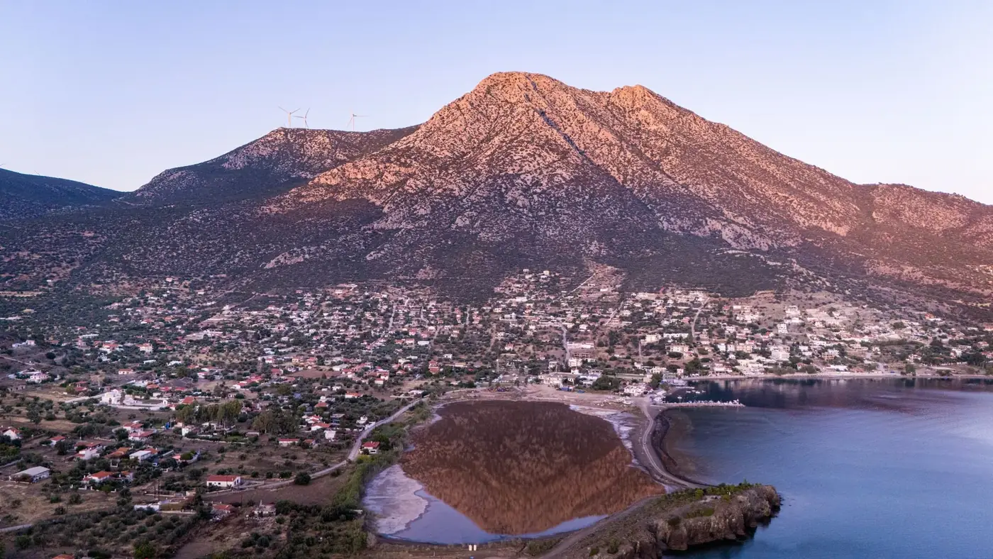 Scenic aerial shot of a Greek mountain village during sunset, with wind turbines and calm waters.