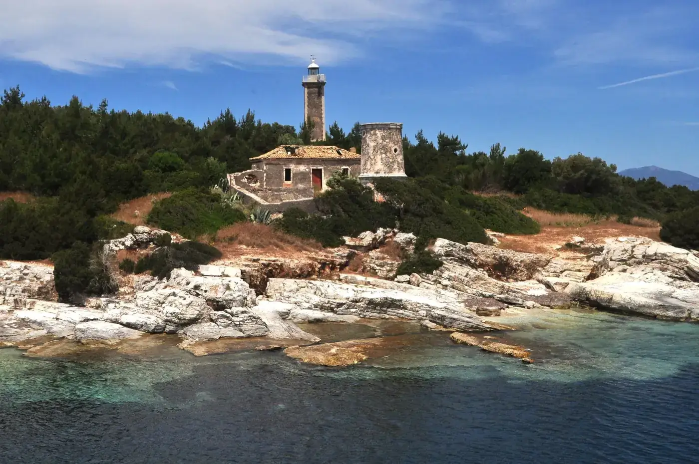 Lighthouse on a rocky coast in Greece, surrounded by clear blue waters and lush greenery.