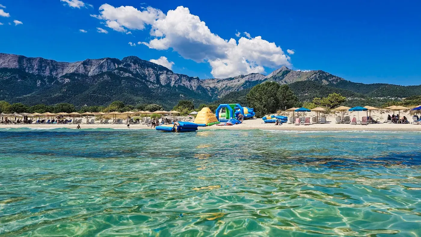 Idyllic beach scene with crystal clear water, mountains, and beachgoers under umbrellas.