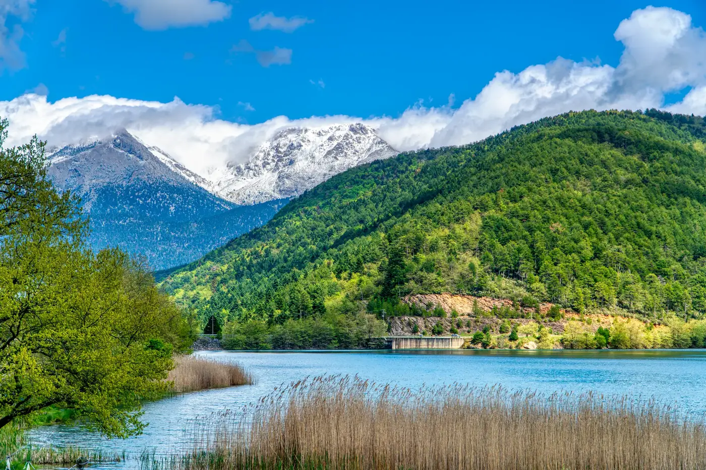 Experience the serene beauty of Lake Doxa surrounded by lush greenery and snow-capped mountains in Greece.