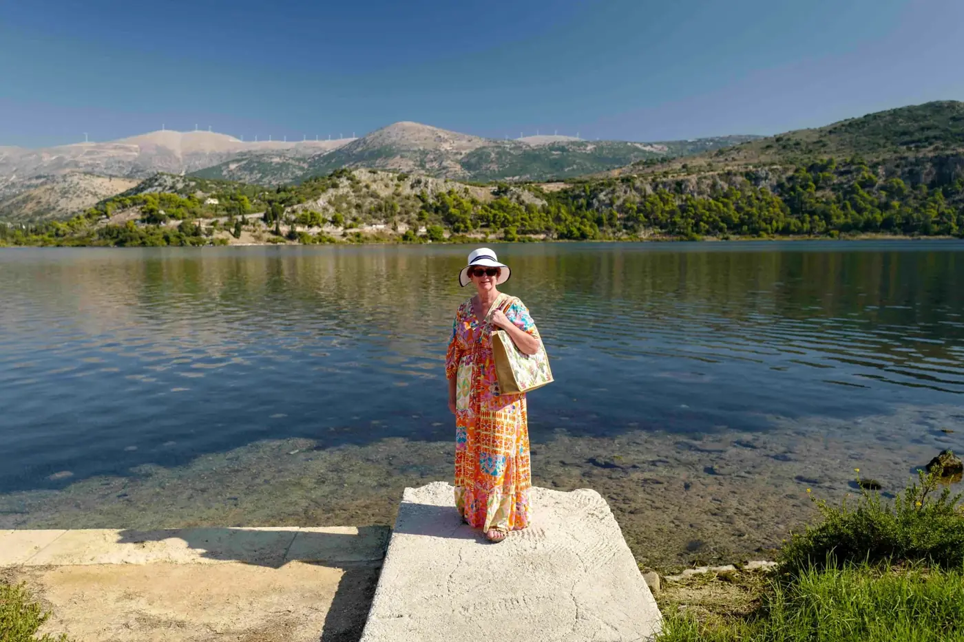 A woman in a bright dress stands by a tranquil Greek lake with scenic hills.